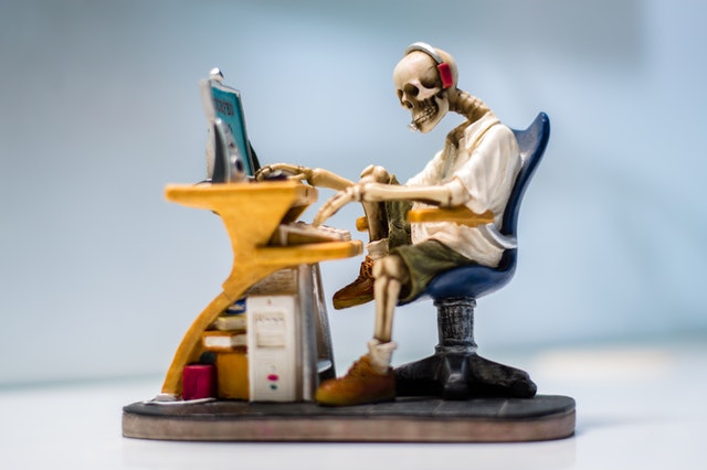Figúrka kostry sedí pri stole a pracuje na počítači